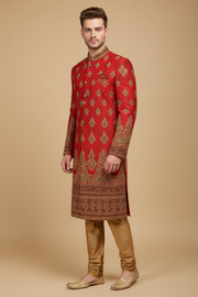 Male model in a red kurta with gold motifs, ornate collar, patterned cuffs and hem, coordinating gold trousers, and tan-gold jutti footwear, front three-quarter view