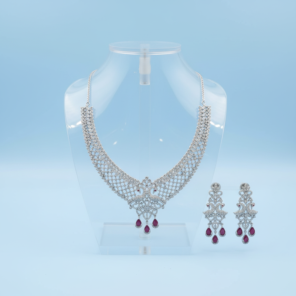 Dhivya silver-tone necklace set with peacock-motif pendant and ruby-red teardrop drops displayed with matching drop earrings on a light blue background
