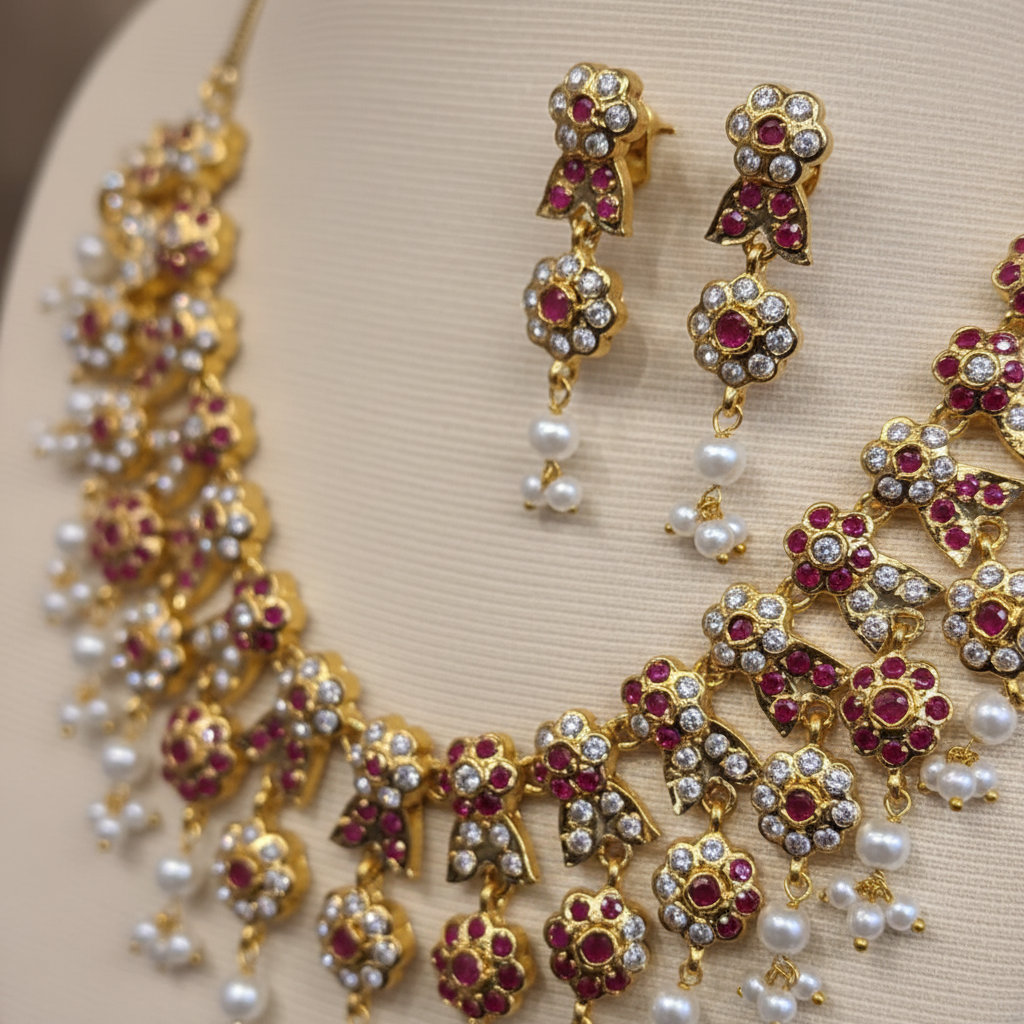 Close-up detail of gold-tone floral necklace and matching earrings with ruby-colored accents, crystal-style stones, and pearl-like drops